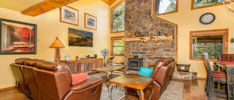 A cozy living room with a stone fireplace, leather furniture, and large windows offering views of the outdoors.