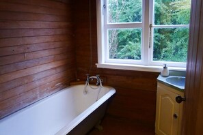 Een badkamer met een houten wand, een vrijstaand bad, een raam met uitzicht op groen, en een klein wastafelmeubel met spiegel.