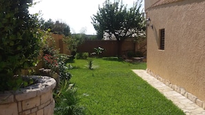 A well-maintained garden with a stone wall, a variety of plants, and a clear pathway leading to a building with a textured wall and a window. - Villetta Orchidea (Giuggianello)