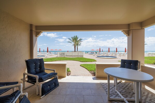 A covered outdoor seating area with a view of a pool and beach umbrellas.