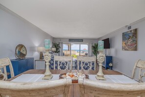 A dining area with a view of the beach, a sign reading 'EAT.BEACH.SLEEP.REPEAT.', and a table set with a basket of fruits.