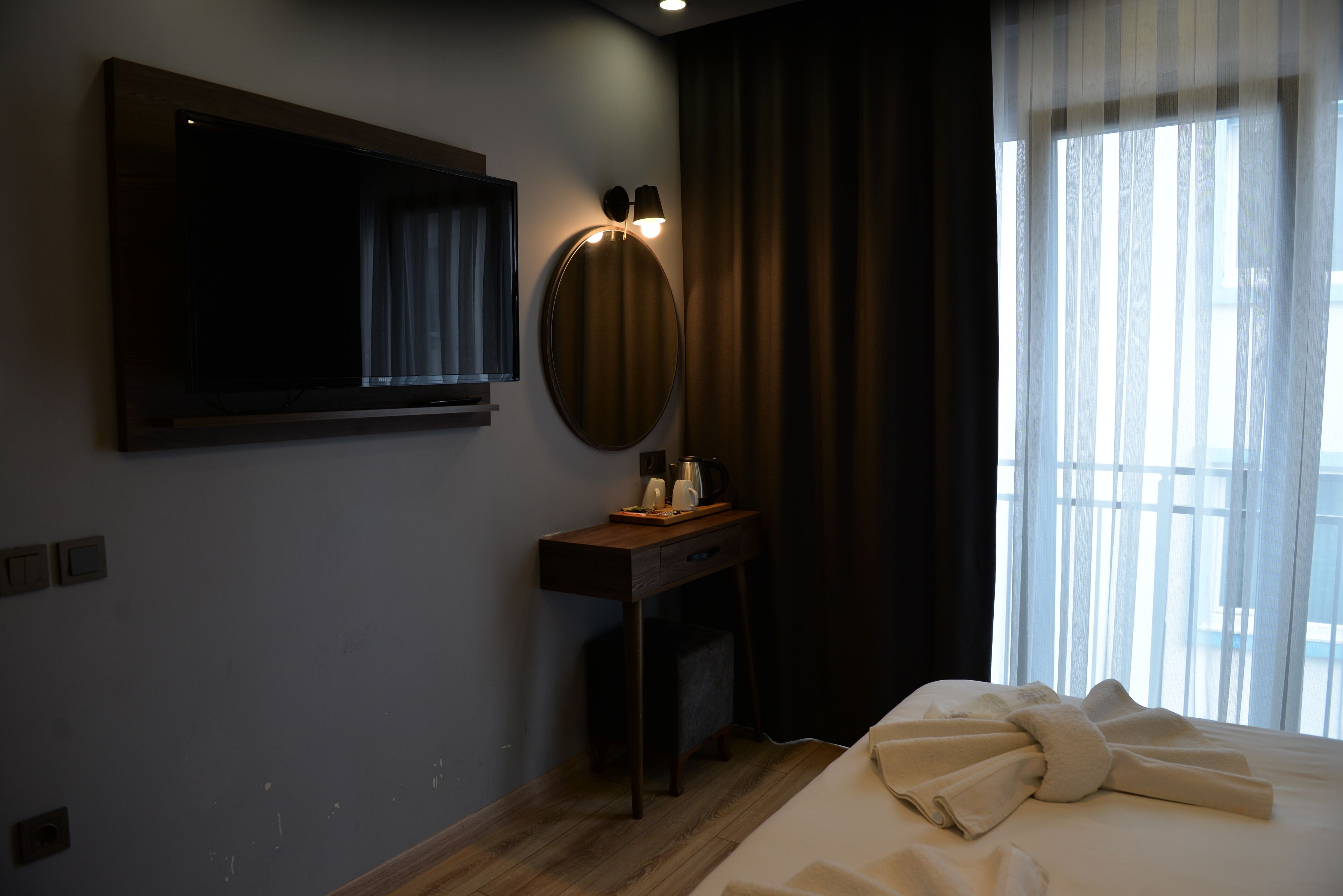 A hotel room with a large bed, a bedside table with a lamp, a TV mounted on the wall, and a window with curtains.