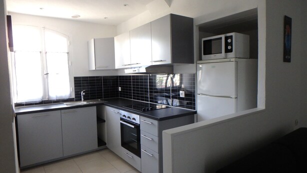 A compact kitchen with grey cabinets, a black countertop, a built-in oven, a microwave, and a window with curtains. - T2 comfortable with aircon (La Londe-Les-Maures)