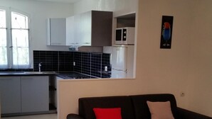 A modern living room with a black sofa, a wooden coffee table, and a red pillow. There is a kitchen area in the background with white cabinets and a black backsplash. - T2 comfortable with aircon (La Londe-Les-Maures)