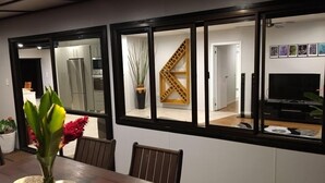 A dining area with a wooden table, chairs, and a vase of flowers. There are large windows with black frames, and a kitchen is visible in the background. - Nevaeh Holiday Home + Pool + Unlimited Internet (Tupapa)