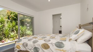 A bedroom with a large window, a bed with patterned bedding, and a view of greenery outside.