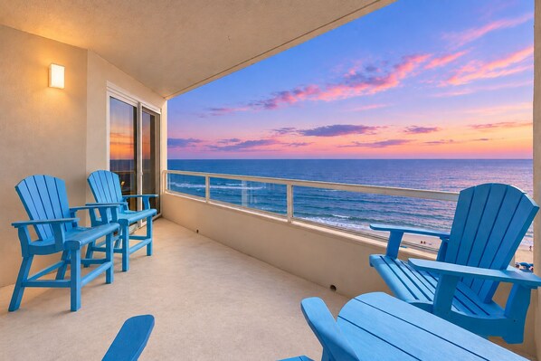 A balcony with a view of the ocean at sunset, featuring blue Adirondack chairs and a glass door.