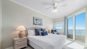 Une chambre avec un grand lit, deux tables de chevet avec des lampes, un ventilateur de plafond, un tableau et une vue sur l’océan par une porte-fenêtre coulissante.