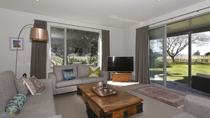 Una sala de estar moderna con un sofá gris, una mesa de centro de madera, un televisor de pantalla plana y ventanas amplias con vistas a un jardín.