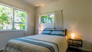 A bedroom with a bed, a nightstand, a lamp, and a window with curtains. - Moa Bach - Blackpool Holiday Home (Oneroa)