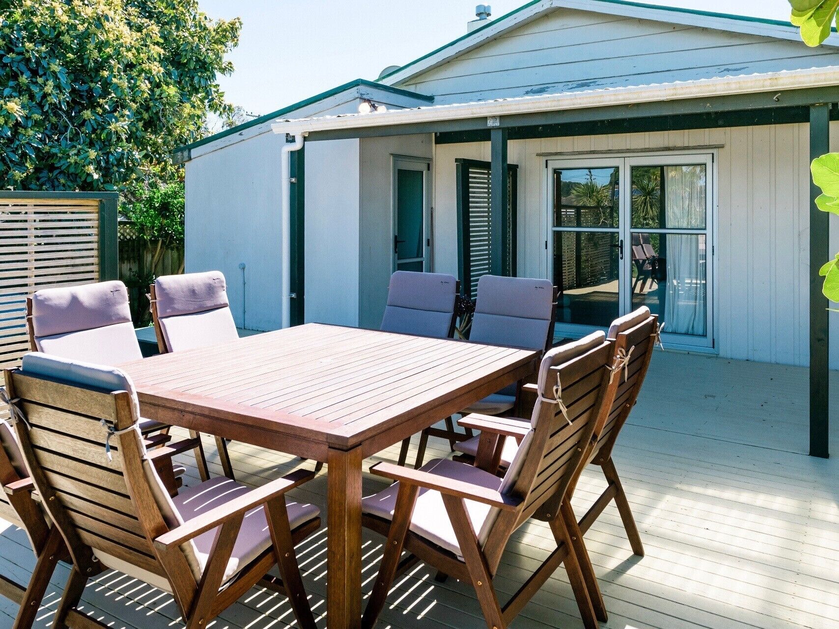 Un patio exterior con mesa y sillas de madera, un espacio de descanso cubierto y una casa al fondo.