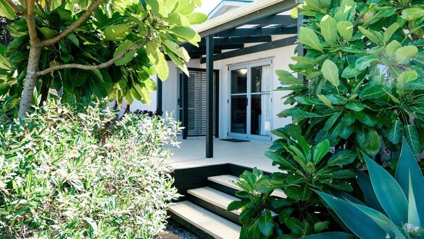 A modern house with a covered porch, surrounded by lush greenery and a gravel pathway. - Moa Bach - Blackpool Holiday Home (Oneroa)