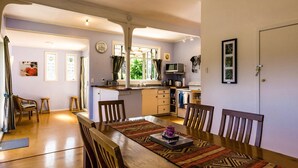 A compact kitchen with a dining area, featuring a wooden table, chairs, and a rug. - Moa Bach - Blackpool Holiday Home (Oneroa)