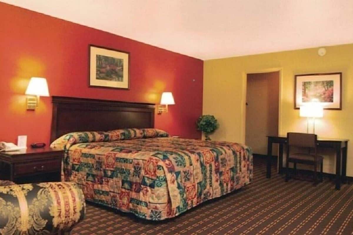 A hotel room with a large bed, two bedside lamps, a desk with a chair, and a door leading to another room.