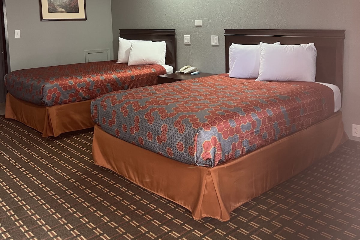 A hotel room with two beds, each with a patterned bedspread and brown skirted base. There is a framed picture on the wall and a telephone on the nightstand.