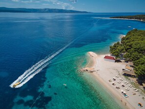 An aerial view of a coastal area with clear blue waters, a sandy beach, and a row of beachside accommodations with umbrellas and lounge chairs.