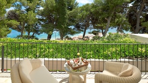 Uma varanda com móveis de vime, uma mesa com uma cesta de frutas e um bule, e vista para o mar e árvores.