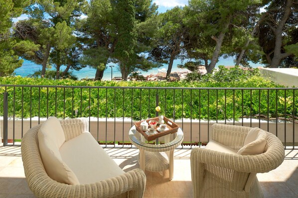 A balcony with wicker furniture, a table with a basket of fruit and a teapot, and a view of the sea and trees.