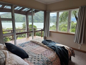 A bedroom with a large bed, a view of a lake, and a balcony with a wooden railing. - Peaceful, private and stunning views (Mahakipawa Arm)
