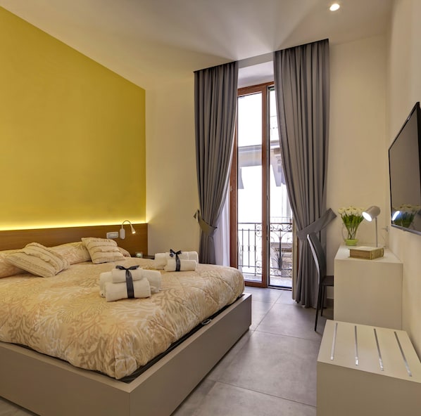 Habitación de hotel moderna con una cama grande, una mesita de noche con lámpara, televisión y un balcón con cortinas.