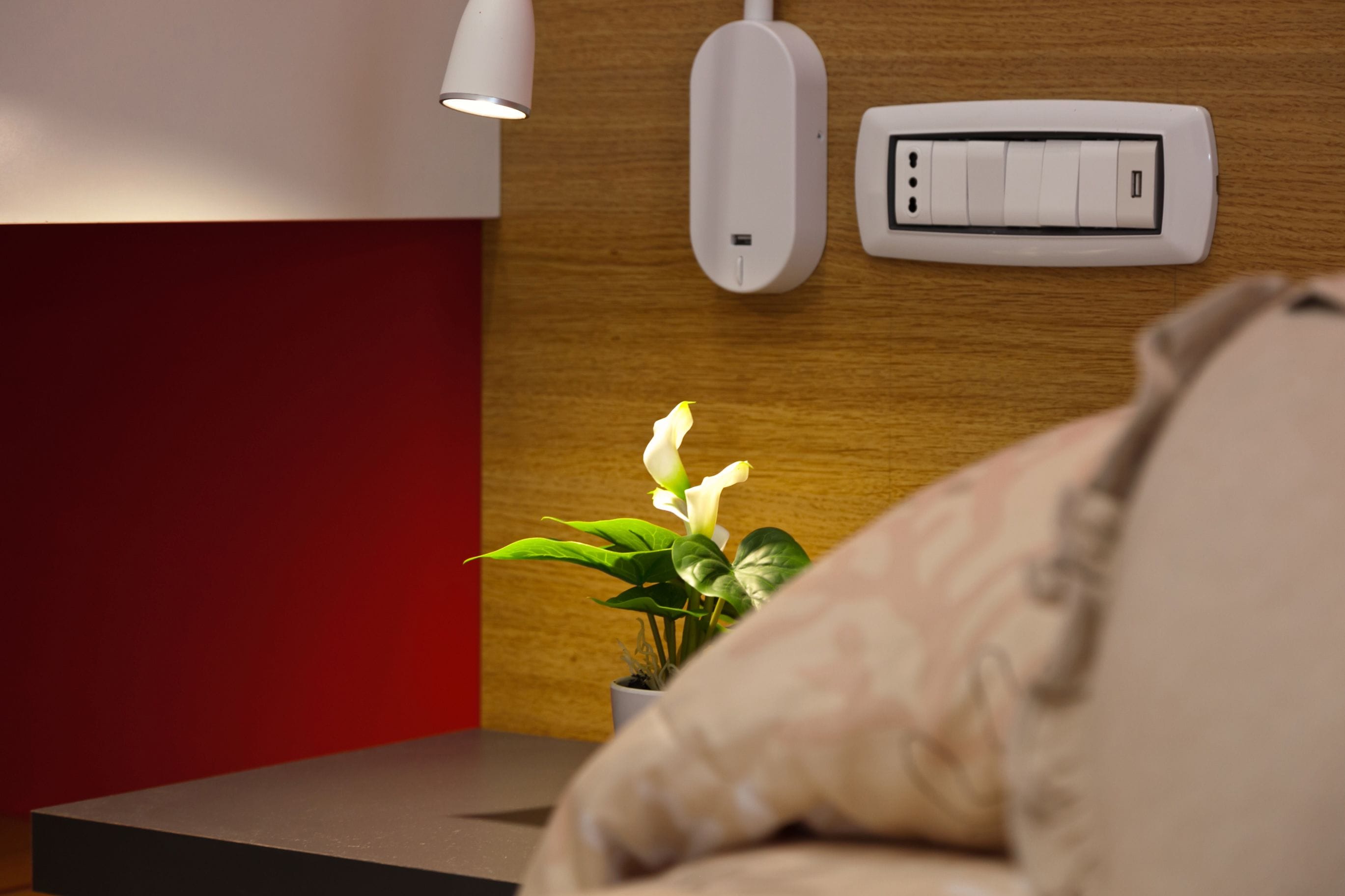 Ein modernes Zimmer mit Nachttisch, Topfpflanze, Wandthermostat und Nachttischlampe.
