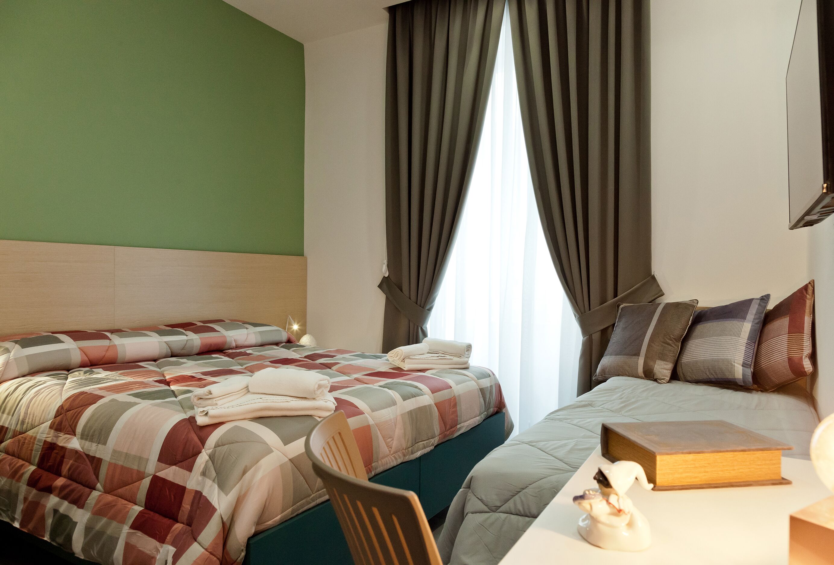 A hotel room with two beds, a green wall, a large window with curtains, and a bedside table with a book and a decorative item.