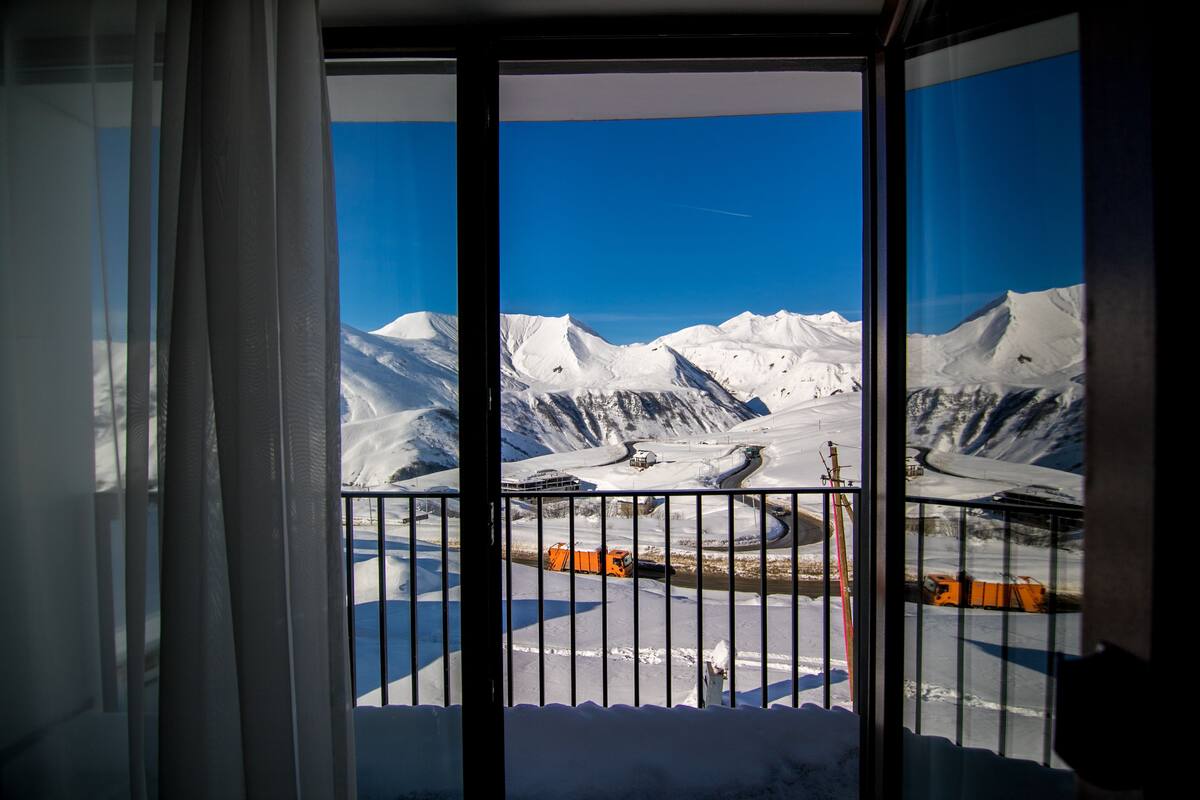 Une vue à travers une fenêtre ornée d’une rambarde en métal, avec en arrière-plan des montagnes enneigées et un ciel bleu dégagé.