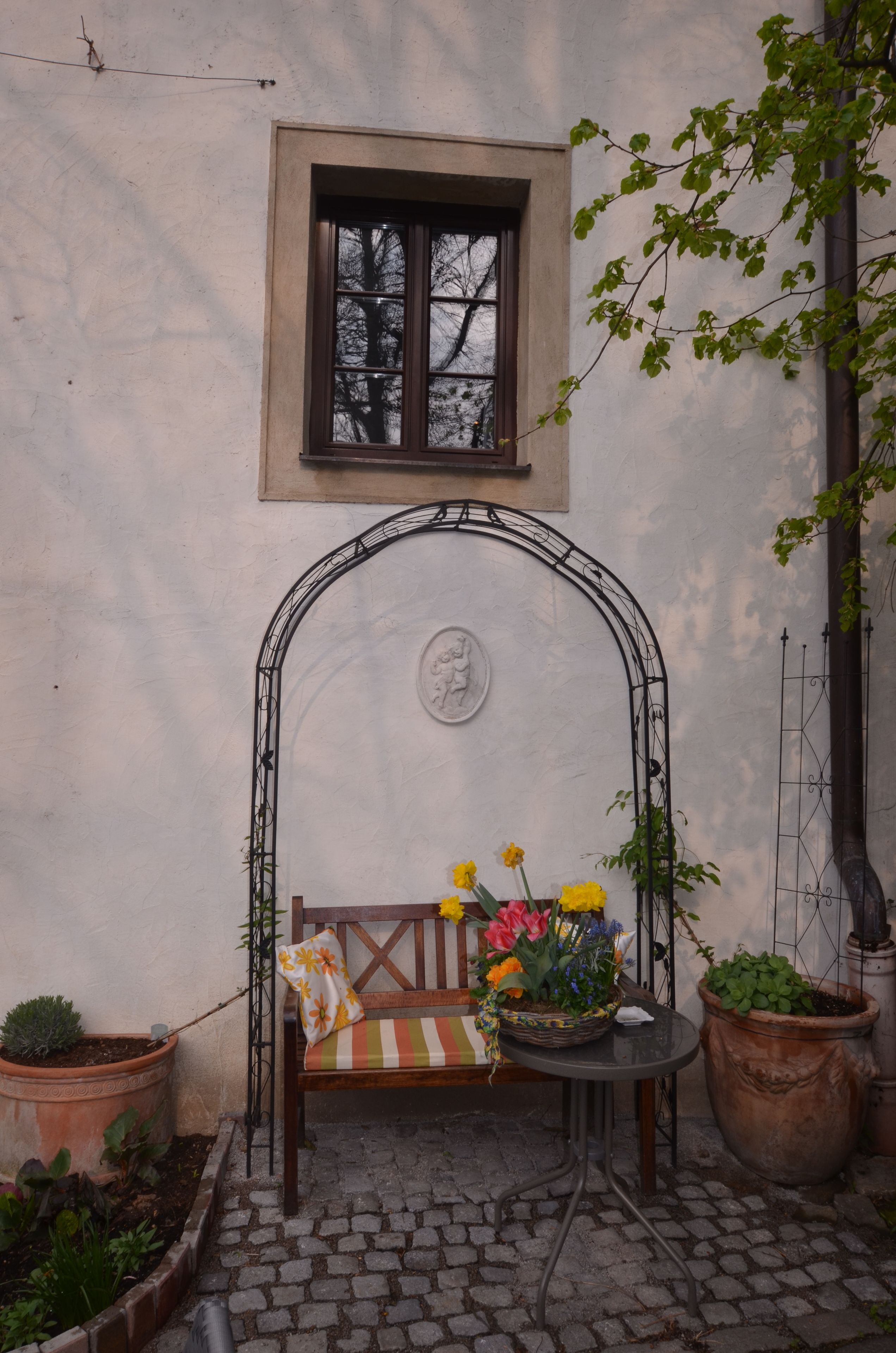 A stone bench with a cushion, a small table with flowers, and potted plants.