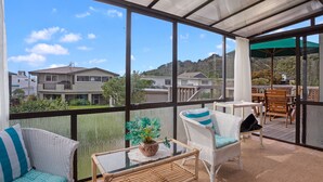 Un espacio al aire libre techado con mobiliario de mimbre, una mesa con una planta en maceta, y vistas a casas y colinas.