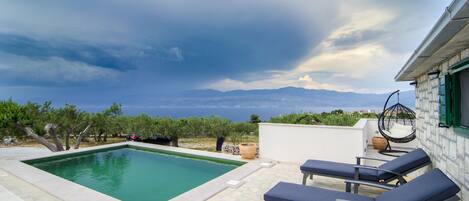 Une piscine entourée de chaises longues et agrémentée d’un fauteuil suspendu, le tout posé sur un sol en pierre et offrant une vue sur la mer et les montagnes.