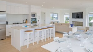 Une cuisine moderne avec des meubles blancs, un îlot central doté de tabourets, un coin repas avec une table pour quatre personnes et un salon avec un canapé et un téléviseur.