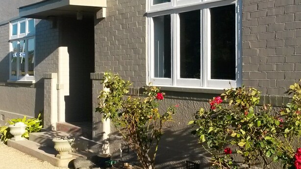 Een gebouw met een gevel van grijze bakstenen, een wit raam en een klein portaal met potplanten en een bloembed.