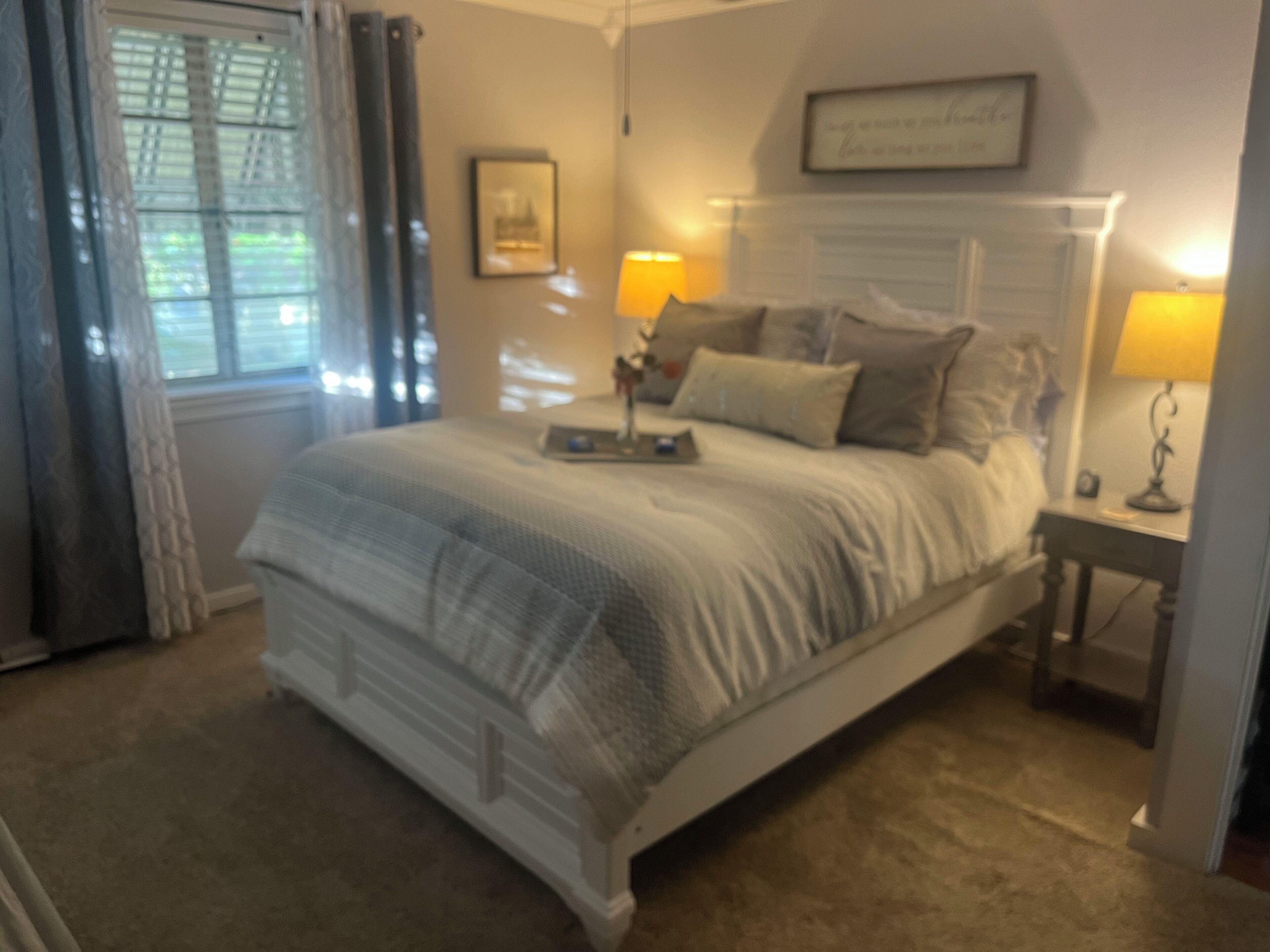 A bedroom with a large bed, two bedside tables with lamps, a window with curtains, and a framed picture on the wall.