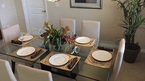 Un coin repas avec une table en verre dressée pour quatre personnes, entourée de chaises blanches. Un pot de fleurs se trouve dans un coin, et un tableau encadré est accroché au mur.