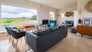 A modern living room with a large window, a grey sofa, a blue armchair, and a TV. - Le Sémaphore - Maison avec sauna Vue mer (Cancale)