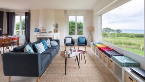 A modern living room with a sofa, chairs, and a dining area. There is a large window with a view of greenery and the sea. - Le Sémaphore - Maison avec sauna Vue mer (Cancale)