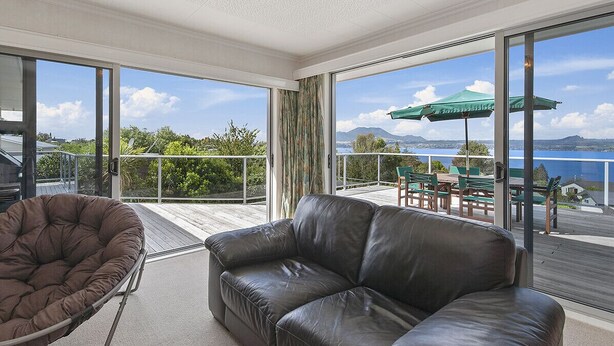 Un salon moderne avec un canapé en cuir et un fauteuil en forme de poire, de grandes fenêtres offrant une vue sur la mer et la verdure, et un balcon aménagé avec du mobilier d’extérieur.
