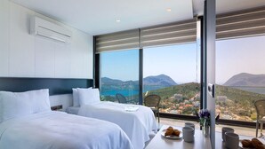 Ein Hotelzimmer mit zwei Betten, einem großen Fenster mit Blick auf das Meer und die Berge sowie einem Tisch mit Frühstücksangeboten.