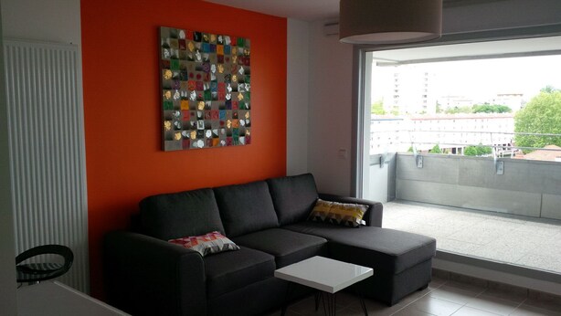 A modern living room with a black sofa, a small white table, and a colorful abstract art piece on the orange wall. - Apartment bayonne T2 (Bayonne)