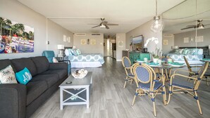 Un vaste espace de vie comprenant un canapé, une table à manger et des chaises, un ventilateur de plafond, et une cuisine à l’arrière-plan.