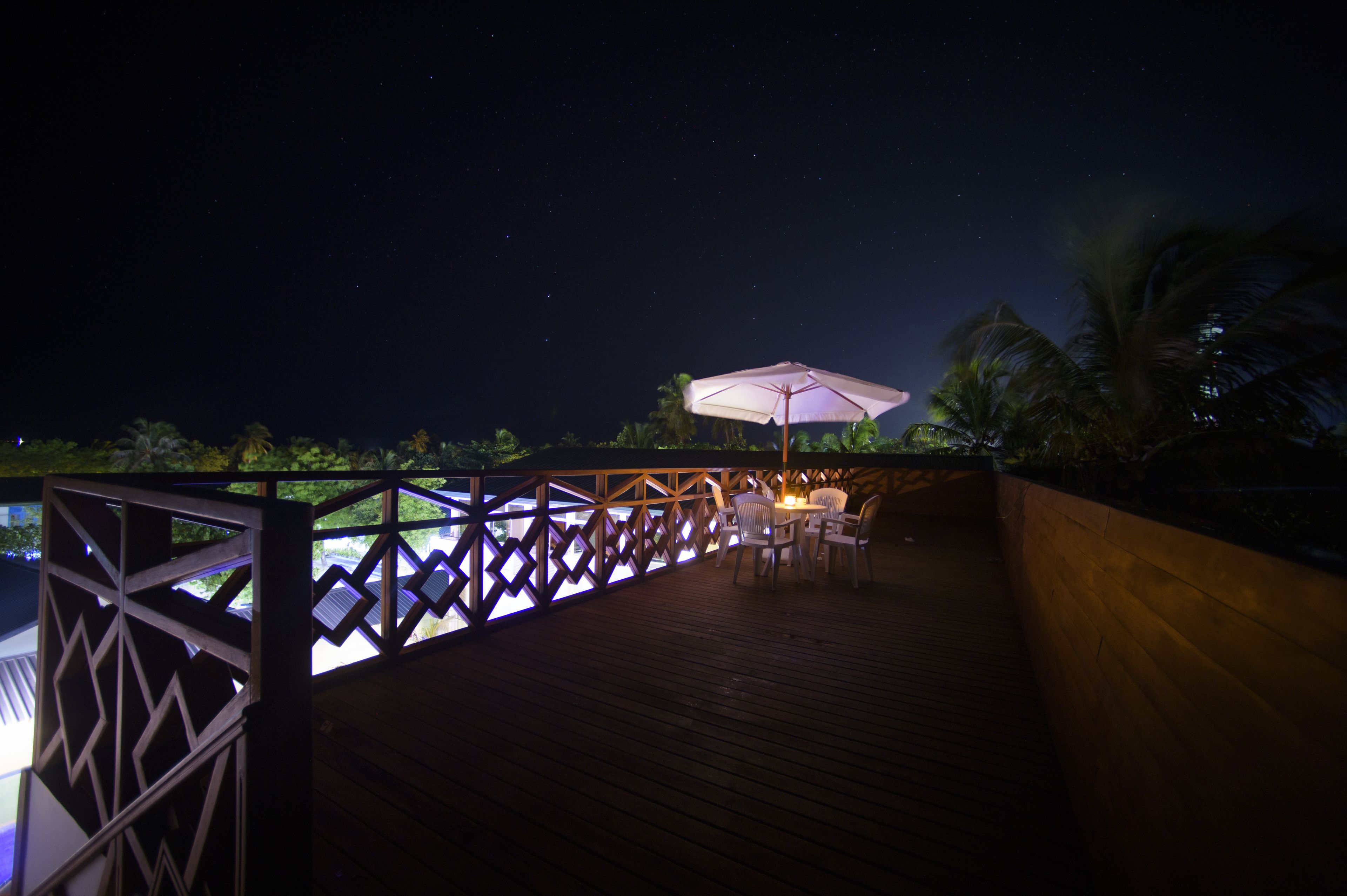 A wooden deck with a table and chairs under a large umbrella, overlooking a pool area at night.