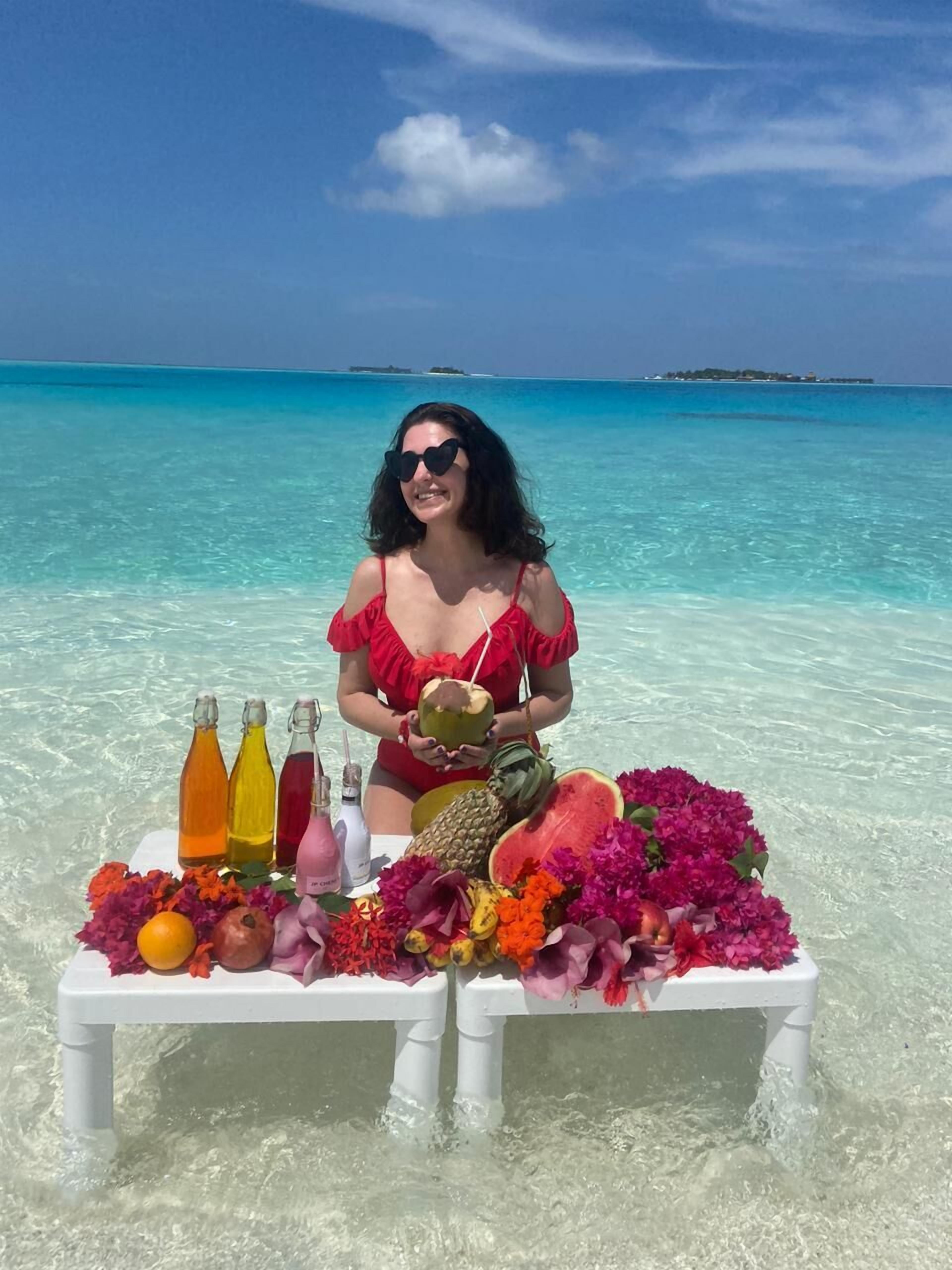 En person sidder på en strand med et bord foran sig, holder en kokosnød. Bordet har flasker, frugt og blomster på.