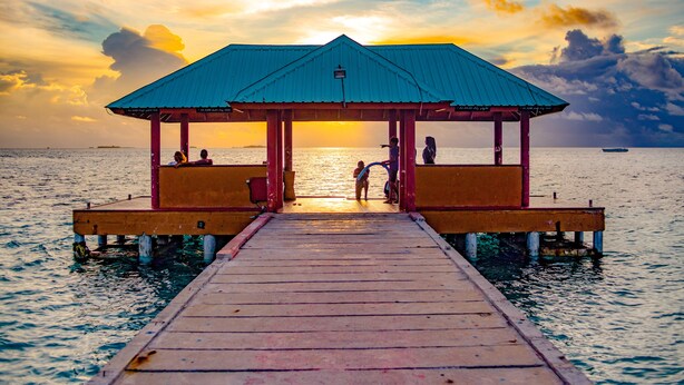 A wooden pier leading to a covered pavilion with a blue roof, situated on a calm body of water. - Perla Dhangethi (Dhangethi)
