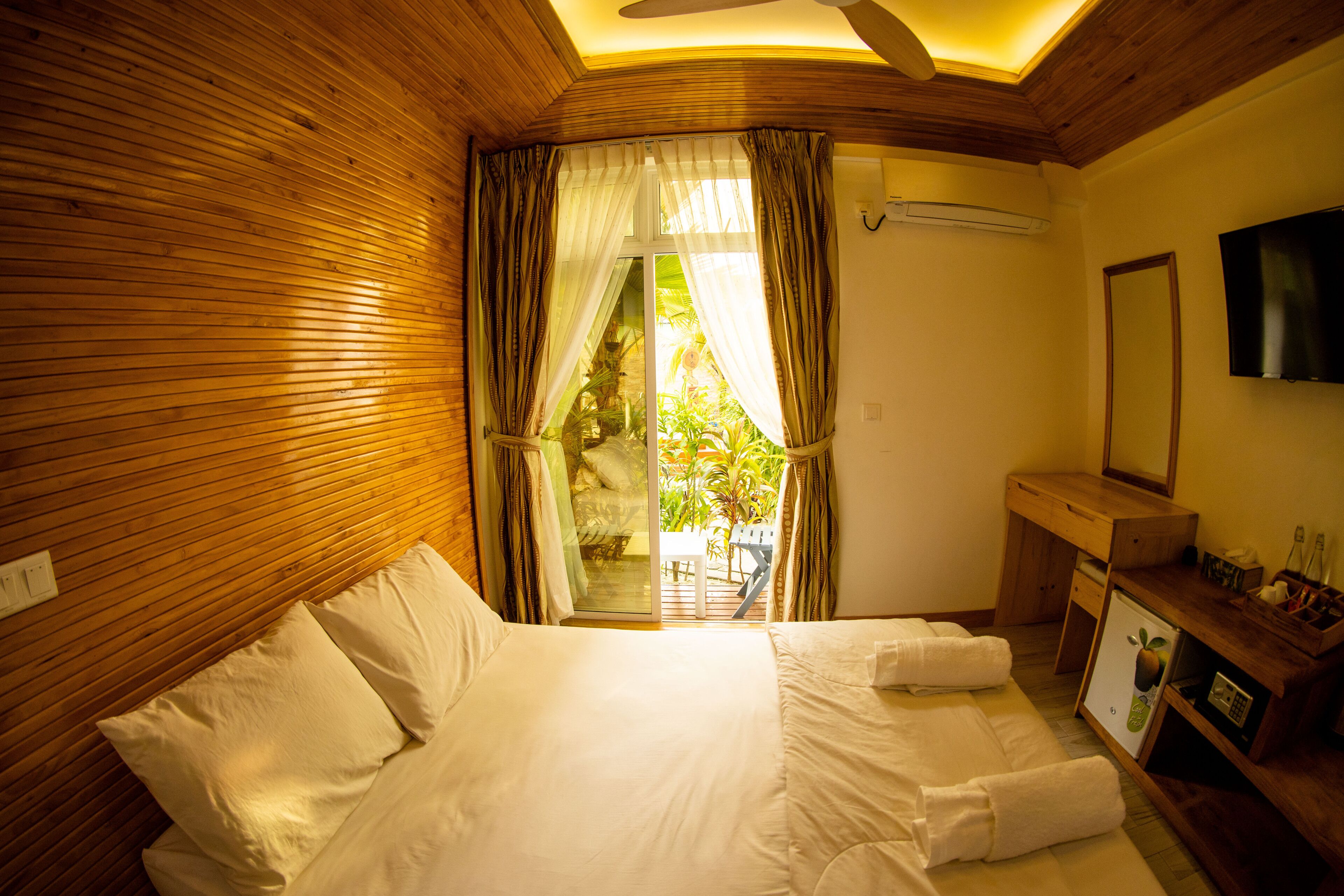 A hotel room with a wooden ceiling, a bed with white linens, a small desk with a mirror, and a TV on the wall.