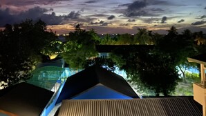A night sky with a visible moon and clouds, a view of a residential area with houses and trees, and a glimpse of a building with a sloped roof. - Perla Dhangethi (Dhangethi)