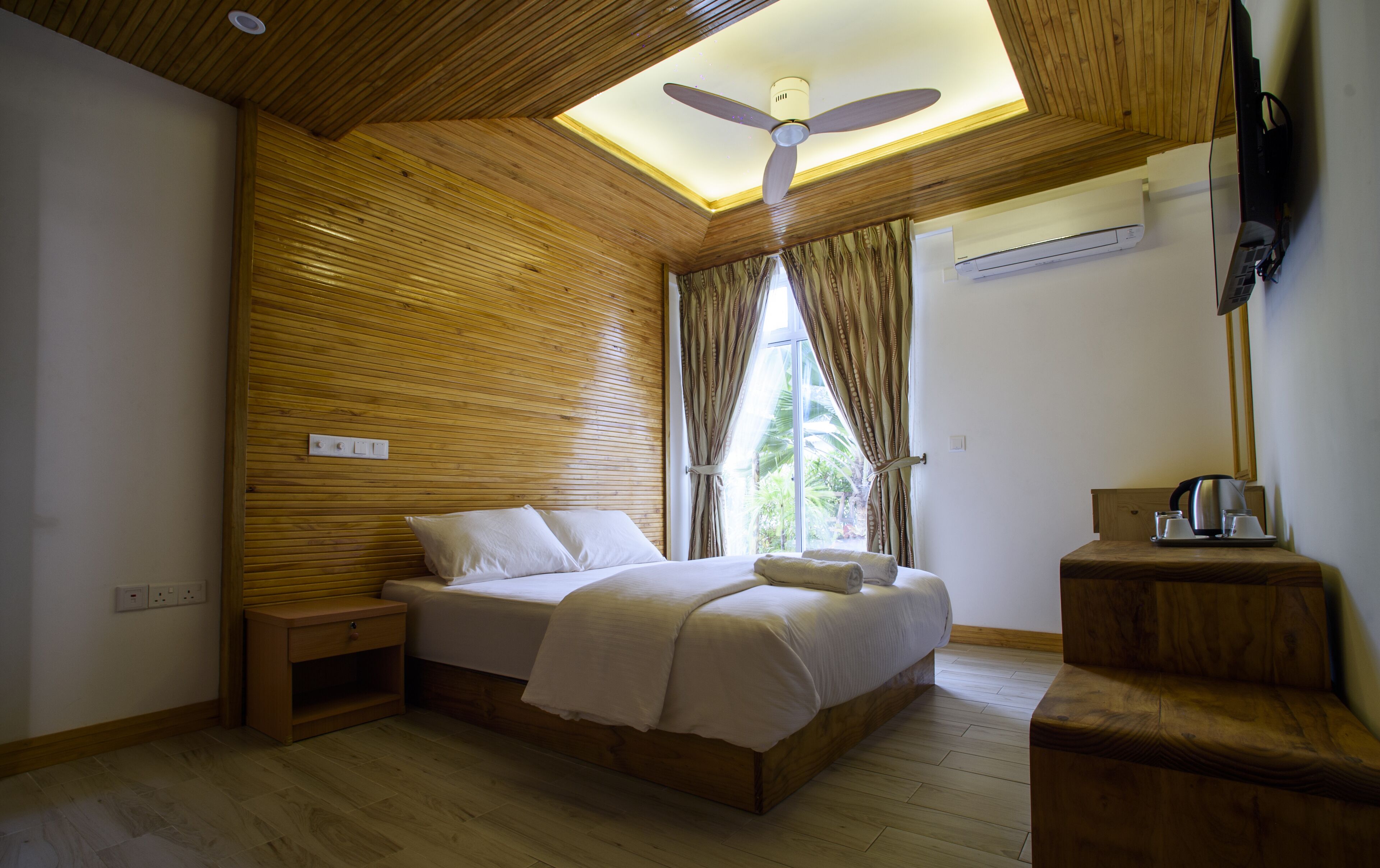 Une chambre avec un plafond en bois, un lit recouvert de draps blancs, une table de chevet, un banc en bois et un ventilateur de plafond.