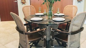 Zona pranzo con tavolo rotondo, quattro sedie in vimini, un vaso con fiori rossi e bicchieri da vino.