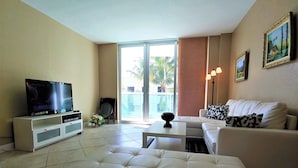 Un soggiorno con una TV su un mobile bianco, un divano bianco con cuscini decorati, un tavolino da caffè e un'ampia porta scorrevole in vetro che dà su un balcone.