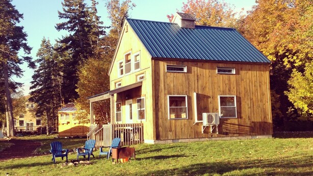 Ein Holzhaus mit Metall-Dach, einer Veranda mit blauen Adirondack-Stühlen und einer Rasenfläche mit Steinrand.