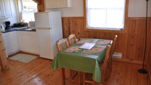 Ein Zimmer mit Holzwandverkleidung, einem Esstisch mit grüner Tischdecke, Holzstühlen, einem weißen Kühlschrank und einem Fenster mit Jalousien.